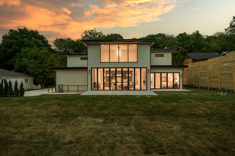 Rear of a modern custom home at dusk, with floor-to-ceiling glass opening onto a stone patio and lawn