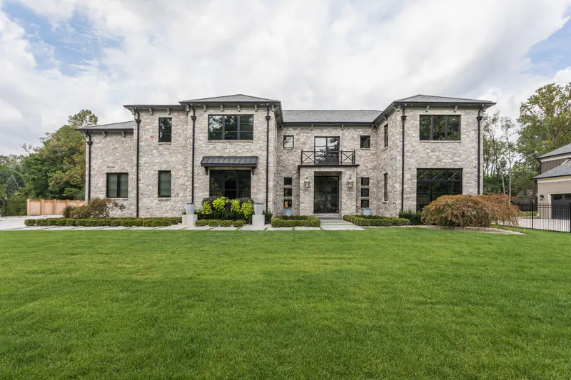 Symmetrical stone estate home with hipped roofs and black windows facing a wide front lawn