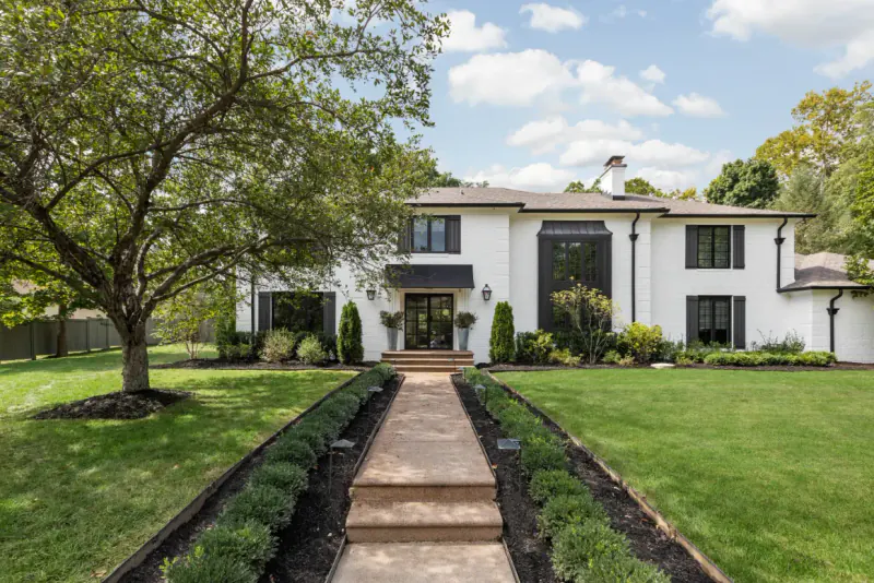 White painted-brick home with black metal awnings and a landscaped front walkway under a shade tree